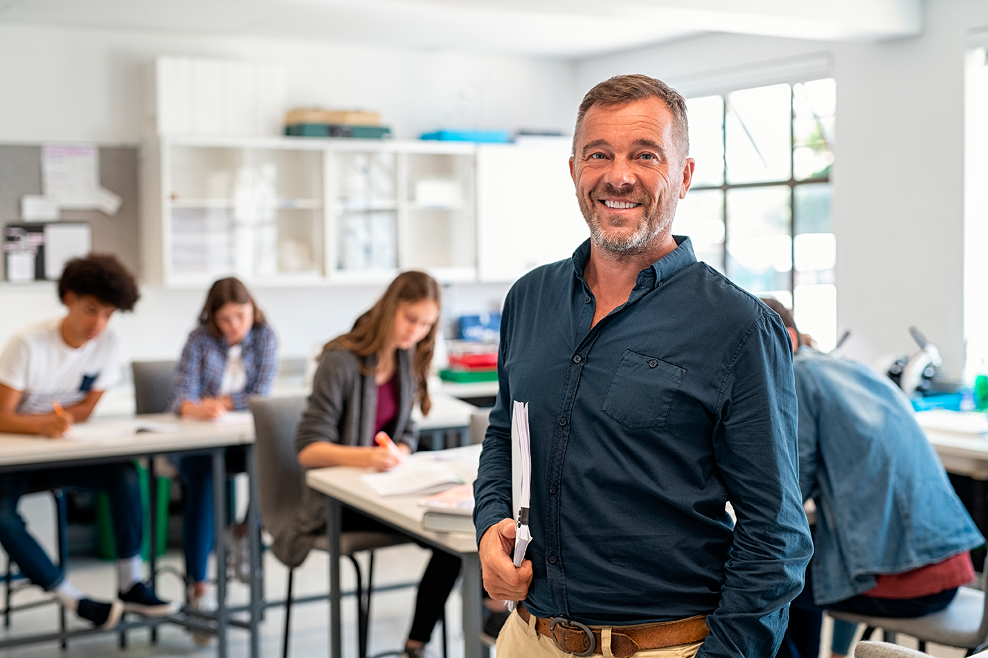 homem com cerca de 40 anos, de camisa social azul escura e livros embaixo do braço, está com sorriso no rosto e atras de dele há alunos em sua escola, a imagem representa este artigo que responde à pergunta É melhor abrir uma escola particular ou adquirir uma franquia de educação pronta?